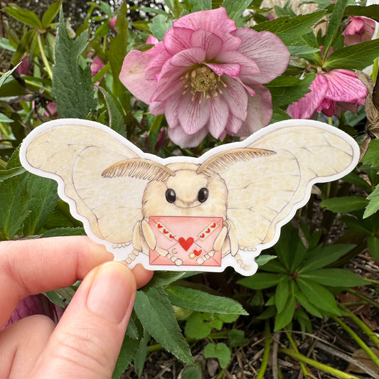 A hand holding a sticker of a tan moth holding a pink love letter.