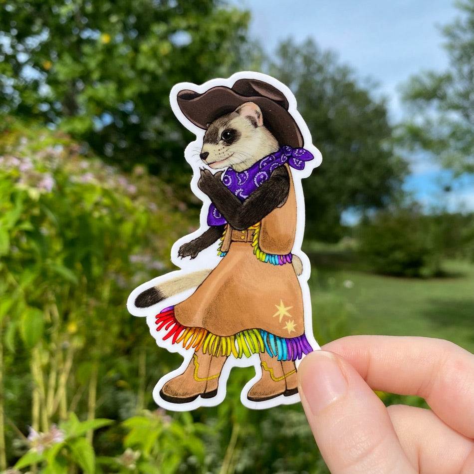 A hand holding a sticker of a black-footed ferret dressed as a cowboy with a pride fringed skirt