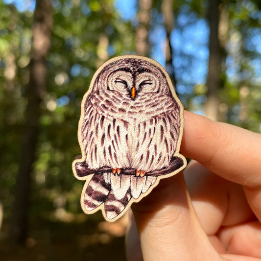 A hand holds a sustainably sourced wooden pin featuring an illustration of a barred owl