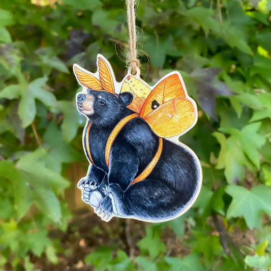 A wooden ornament strung with twine of an American black bear with io moth wings and antennae.