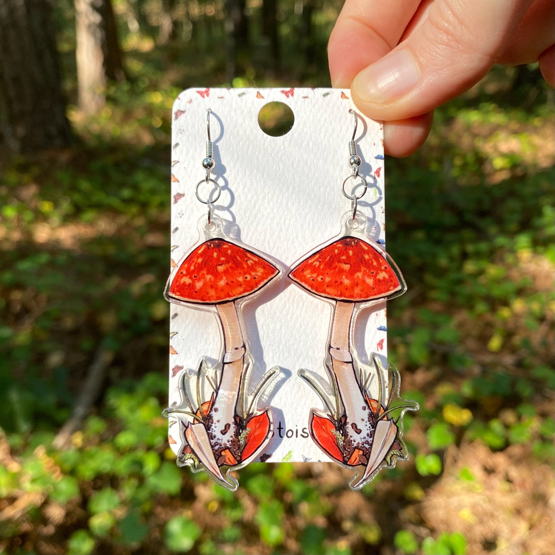 A hand holding a pair of acrylic earrings of amanitas