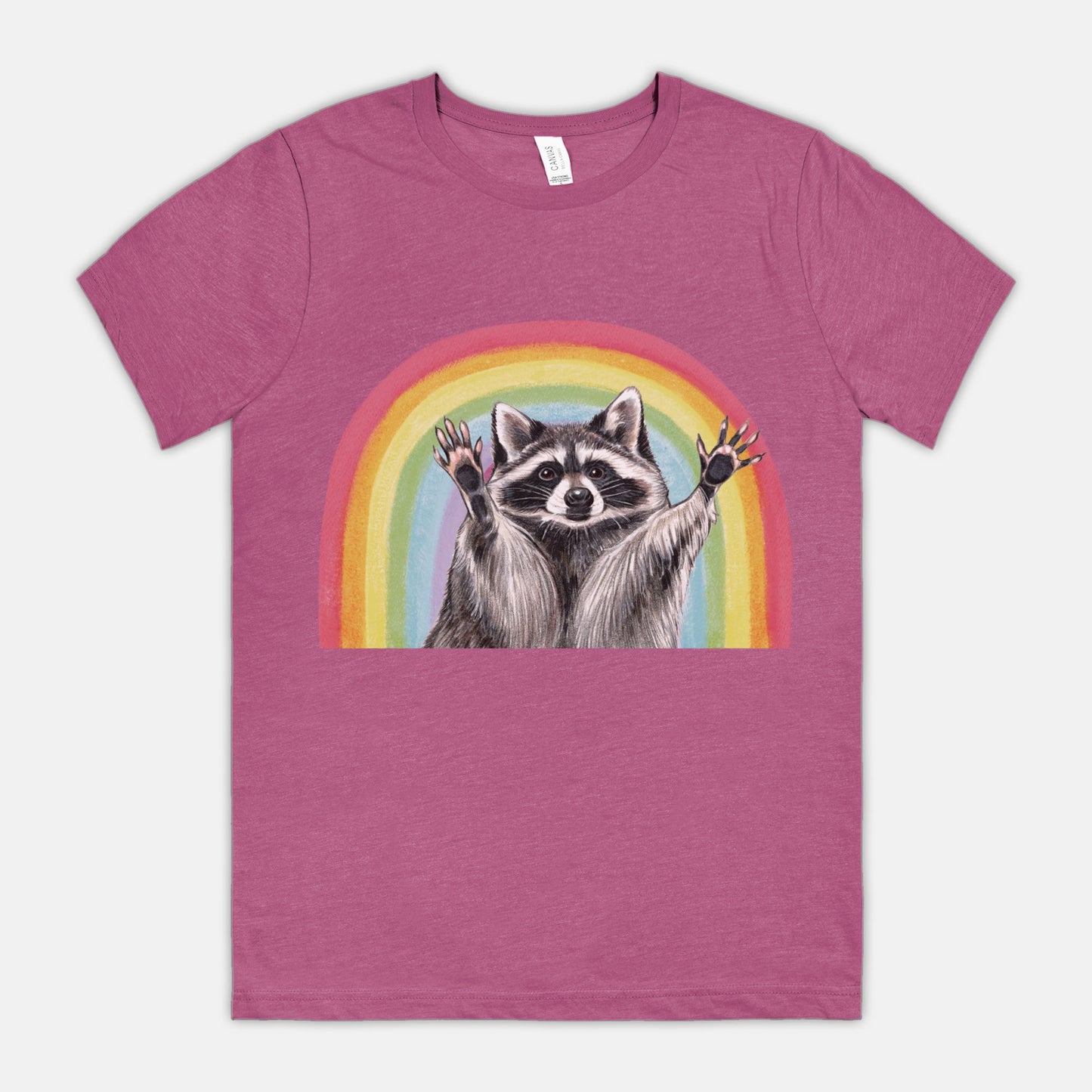 A magenta-colored t-shirt featuring an illustration of a raccoon with raised hands in front of a rainbow.