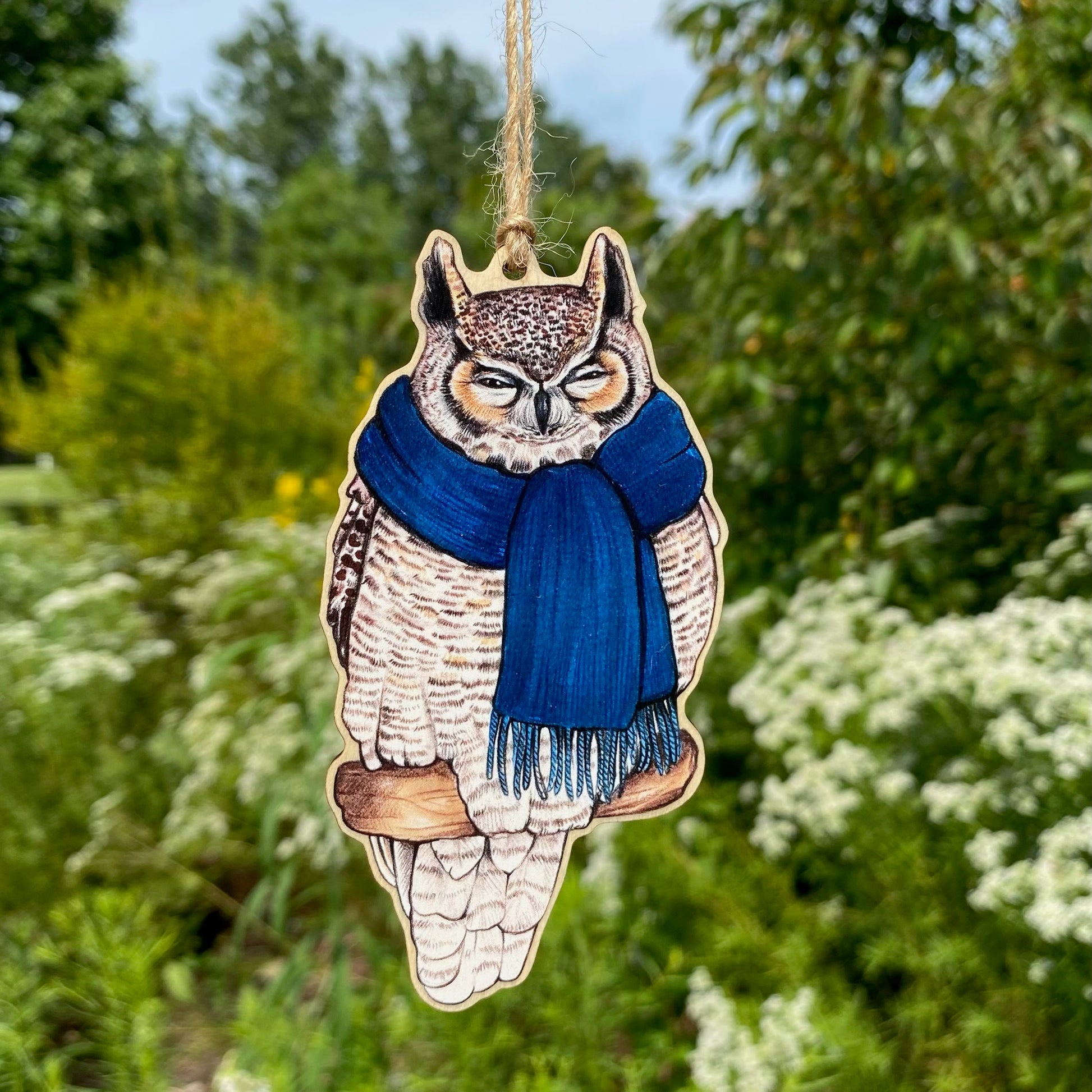 A wooden ornament strung with twine of a great horned owl in a blue scarf.