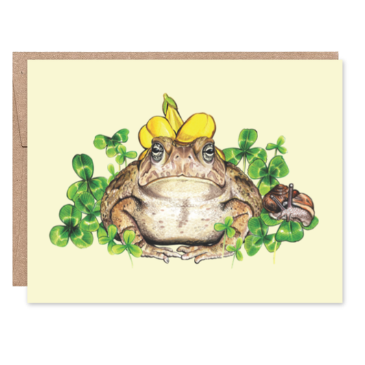 Card with a frog wearing a flower hat surrounded by clovers on a yellow background