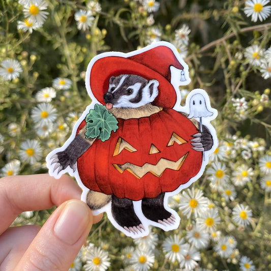 A hand holding a weatherproof vinyl sticker of an American badger in a red pumpkin and witch hat and holding a little cake pop shaped like a ghost.