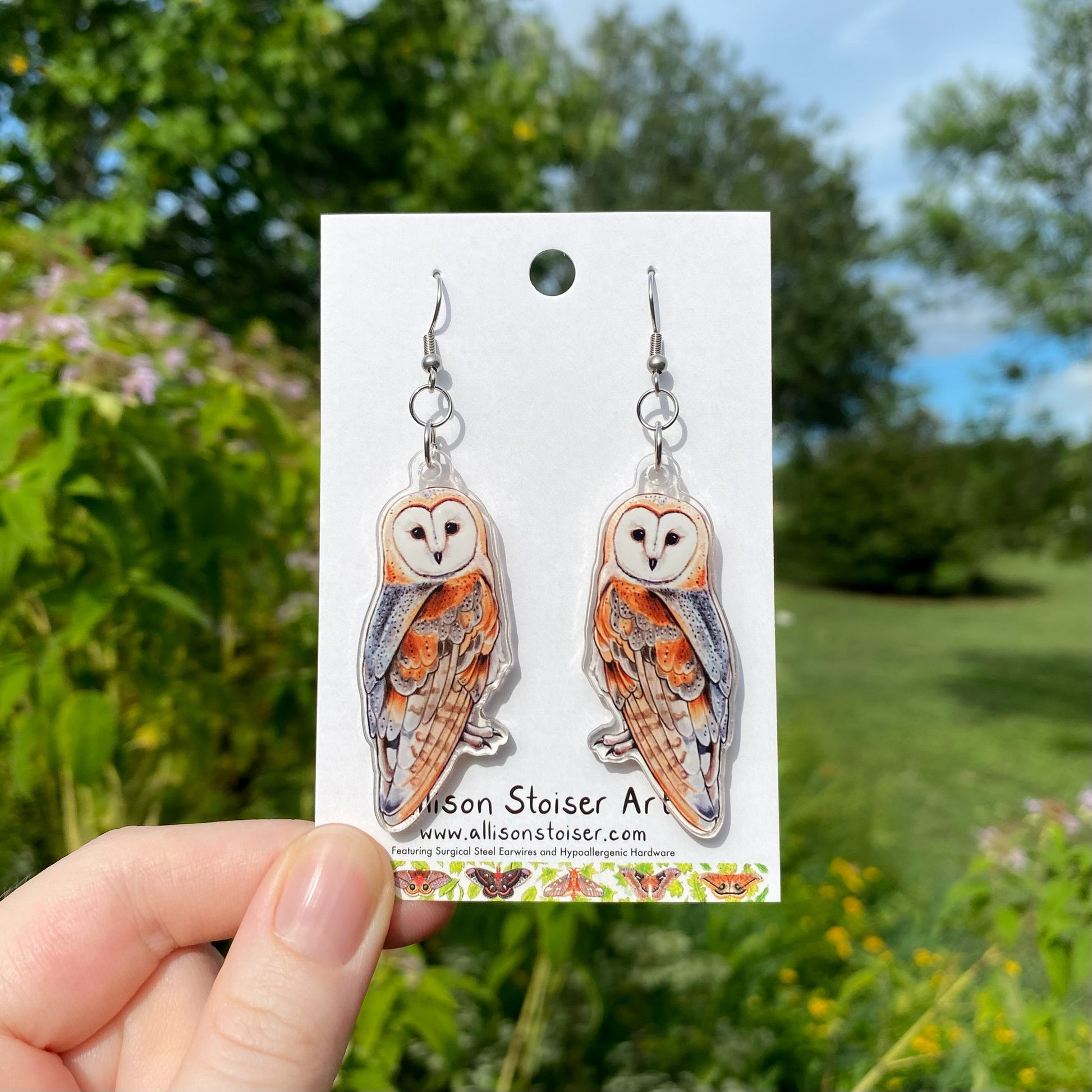 A hand holding a pair of acrylic earrings of barn owls.