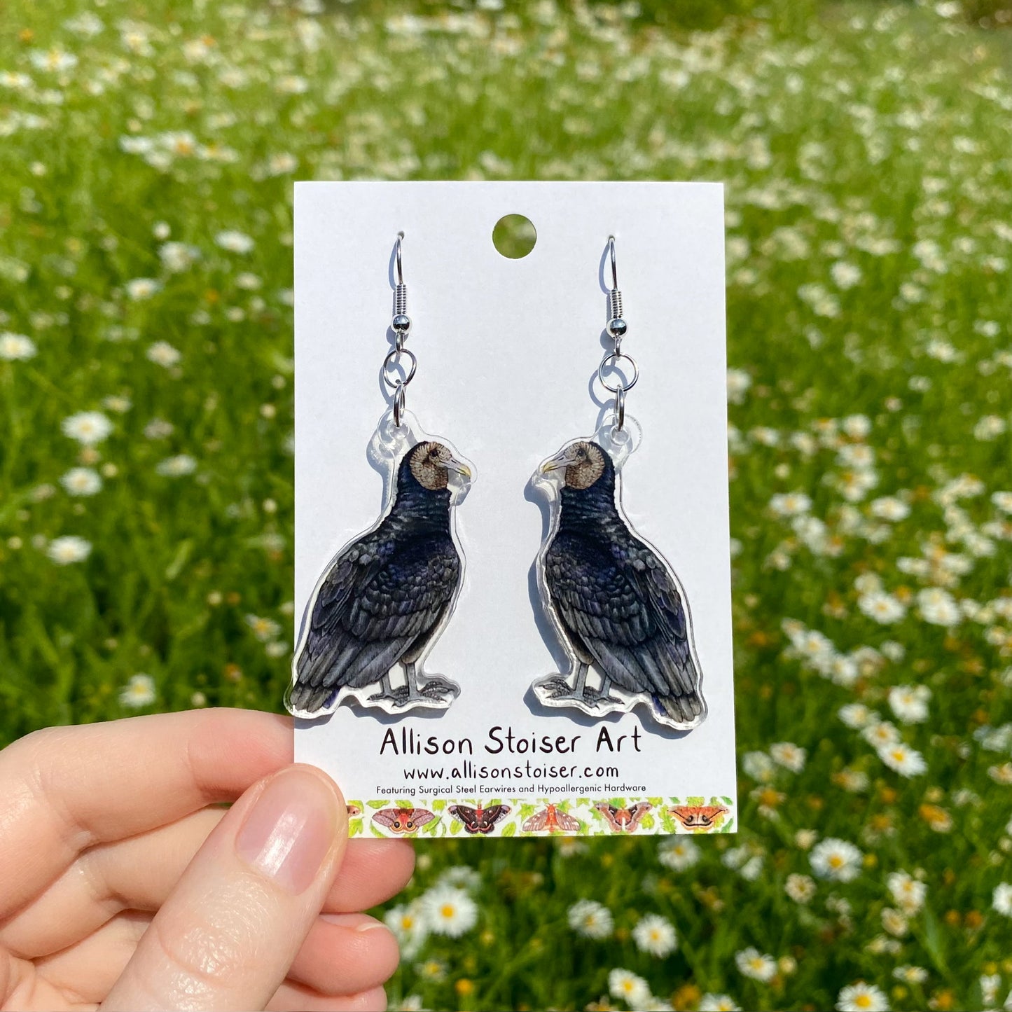 A hand holding a pair of clear acrylic earrings of black vultures.