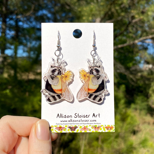 A pair of acrylic earrings of a raccoon wearing a bumblebee costume.