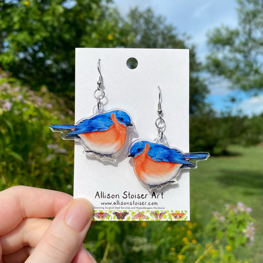 A hand holding a pair of acrylic earrings of eastern bluebirds.