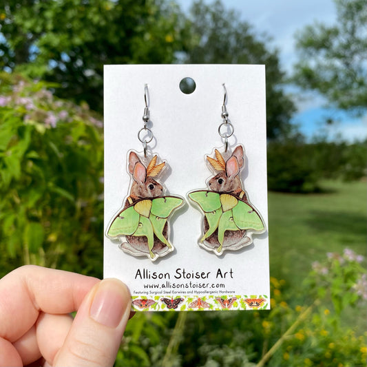 A hand holding a pair of acrylic earrings of an eastern cottontail rabbit wearing luna moth wings