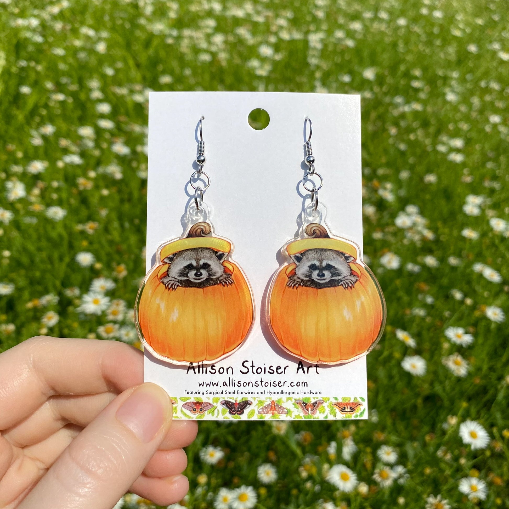 A hand holding a pair of clear acrylic earrings of a raccoon in a pumpkin.