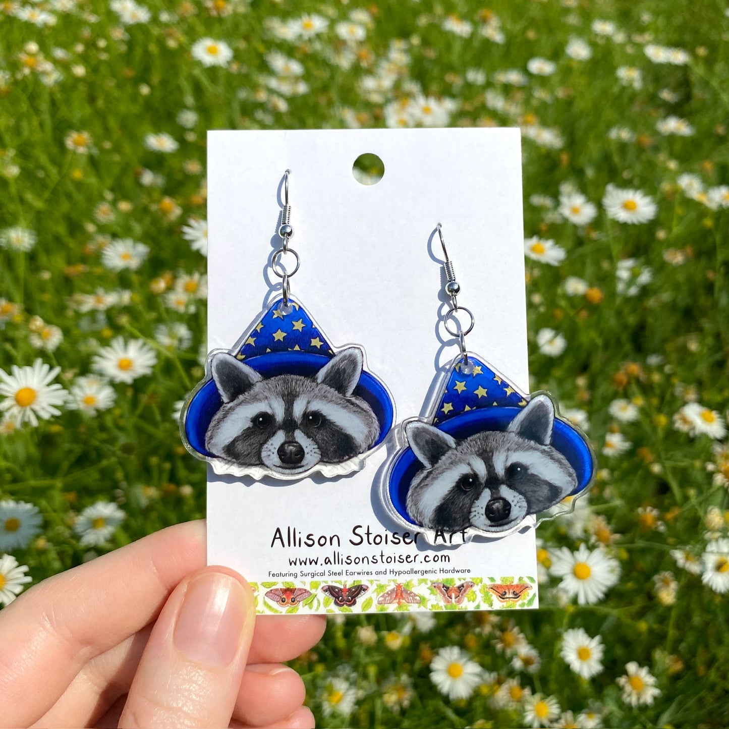 A hand holding a pair of clear acrylic earrings of raccoon in blue wizard hats with stars.