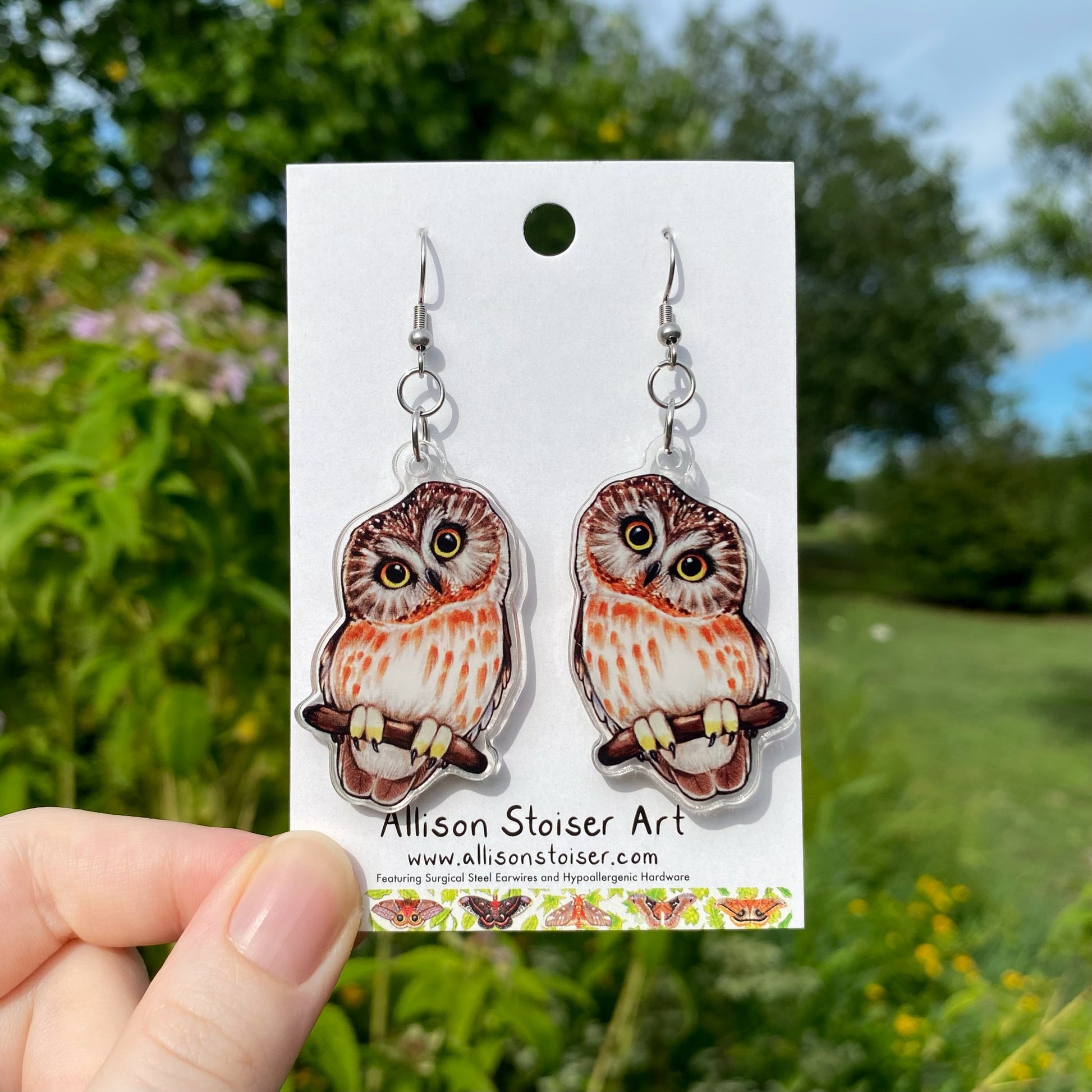 A hand holding a pair of acrylic earrings of saw-whet owls.