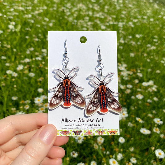 A hand holding a pair of clear acrylic earrings of orange tiger moths.