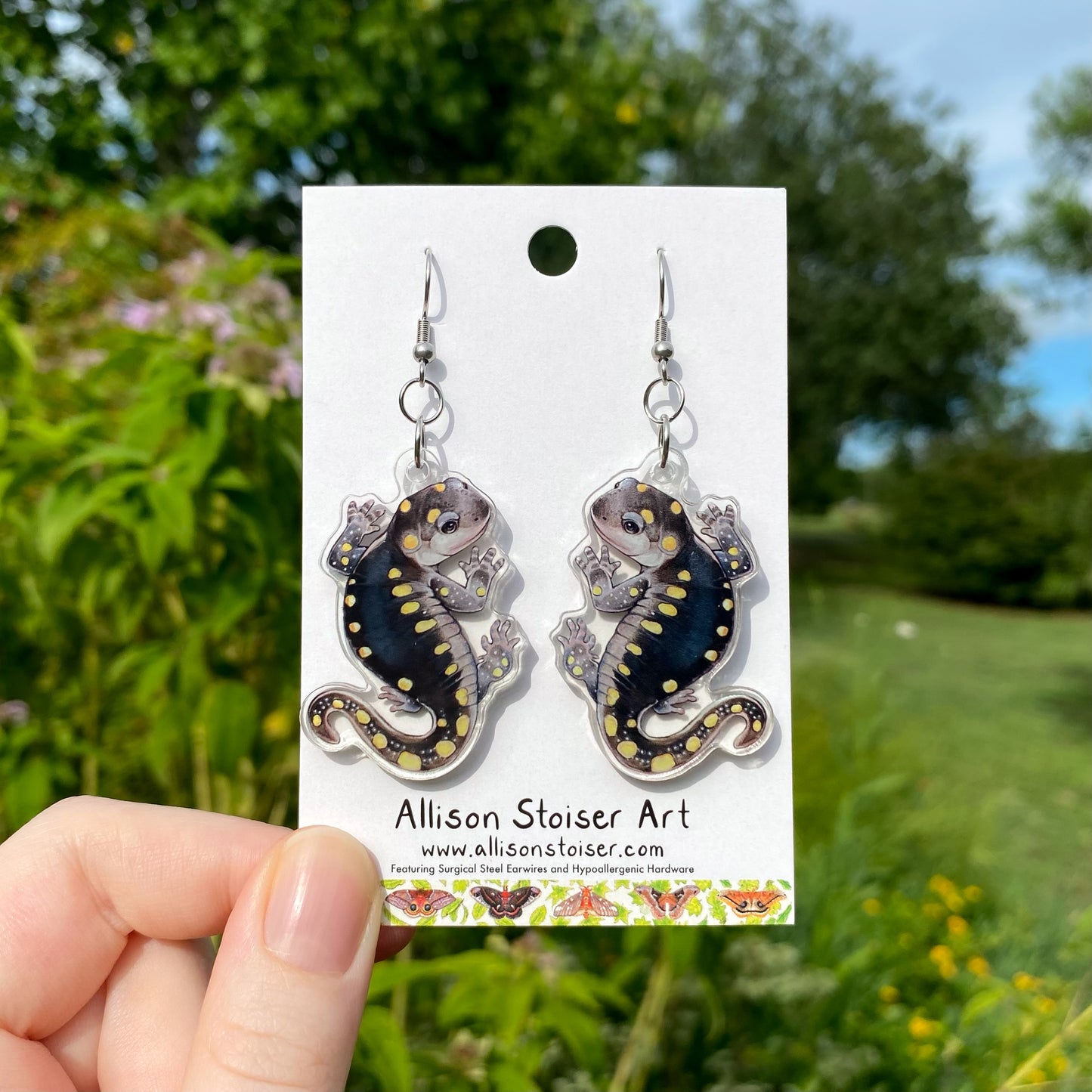 A hand holding a pair of acrylic earrings of yellow-spotted salamanders.