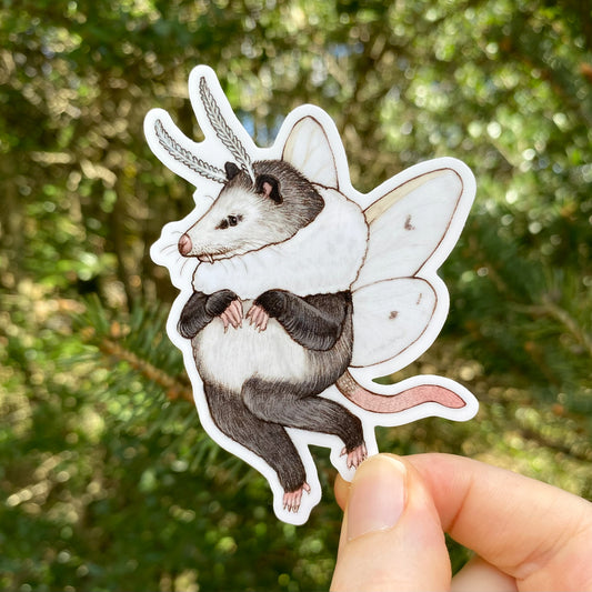 A hand holding a sticker of an opossum with white tiger moth wings. 