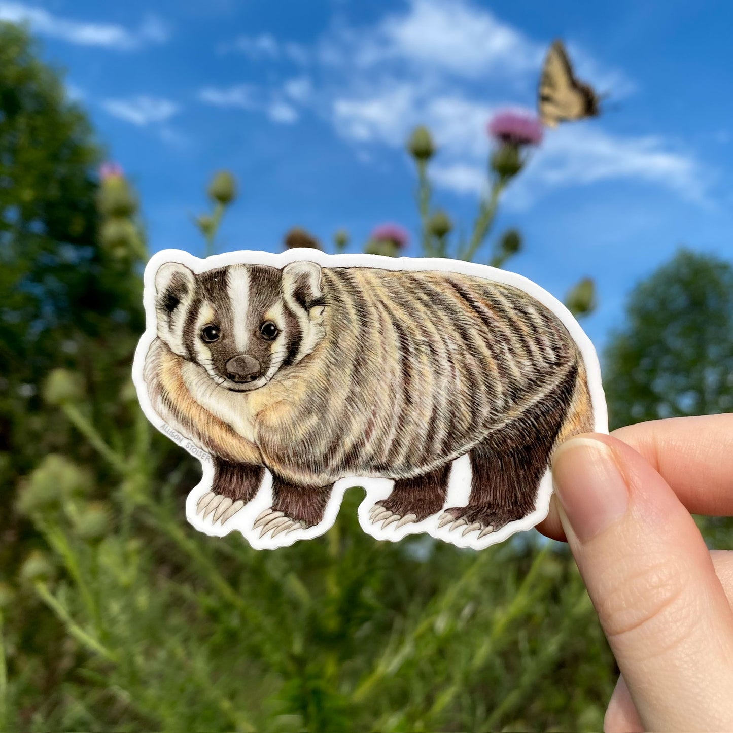 Sticker of a badger held in front of a natural background with plants and a butterfly.