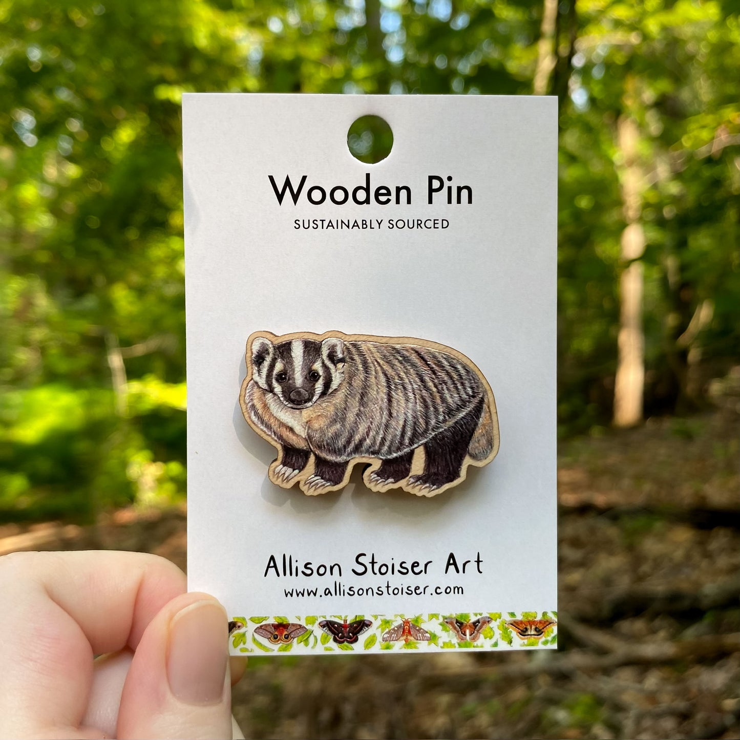 Wooden pin of a badger on a card with 'Allison Stoiser Art' branding, held against a natural background.