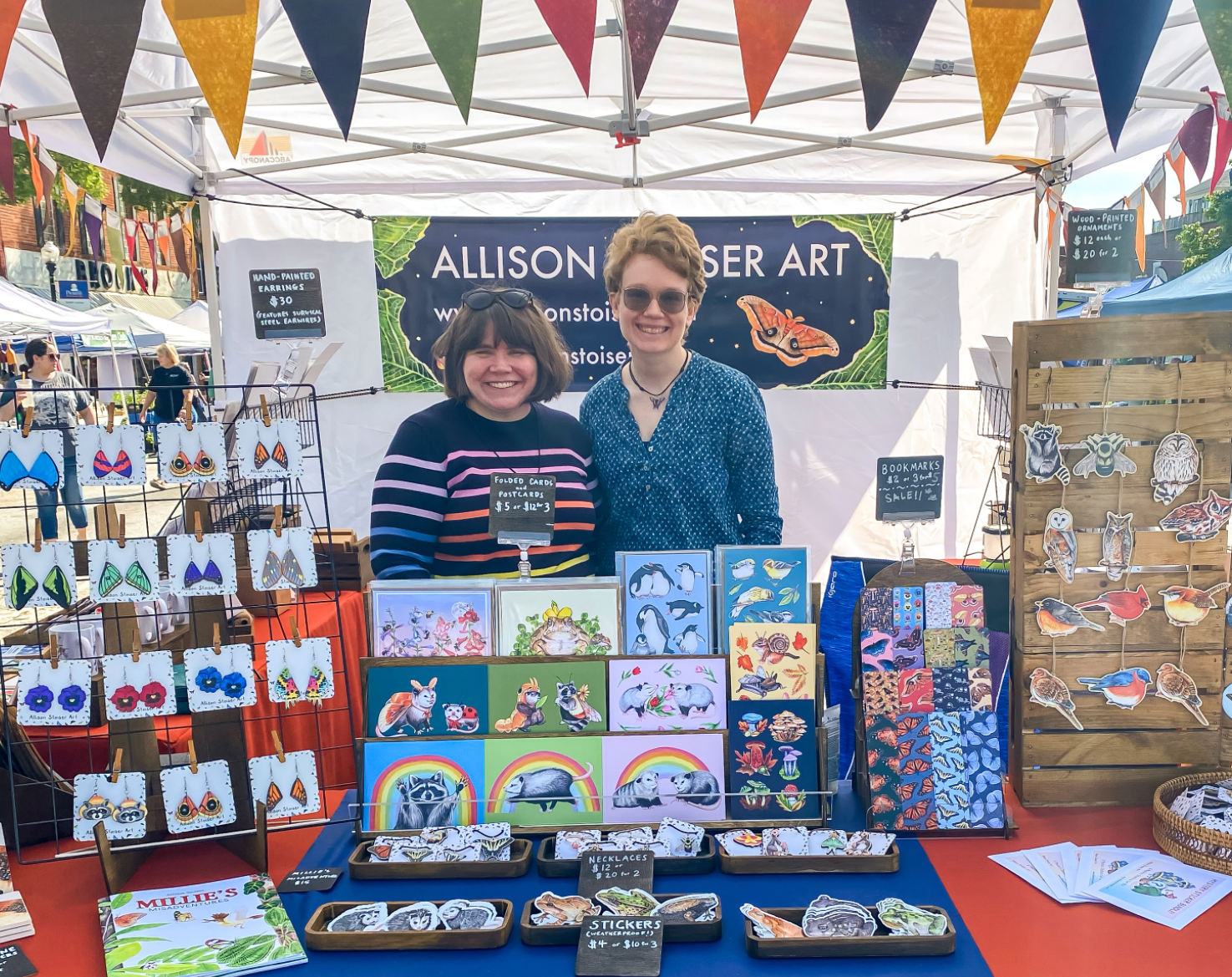 Two women pose proudly in their craft fair booth that showcases nature art