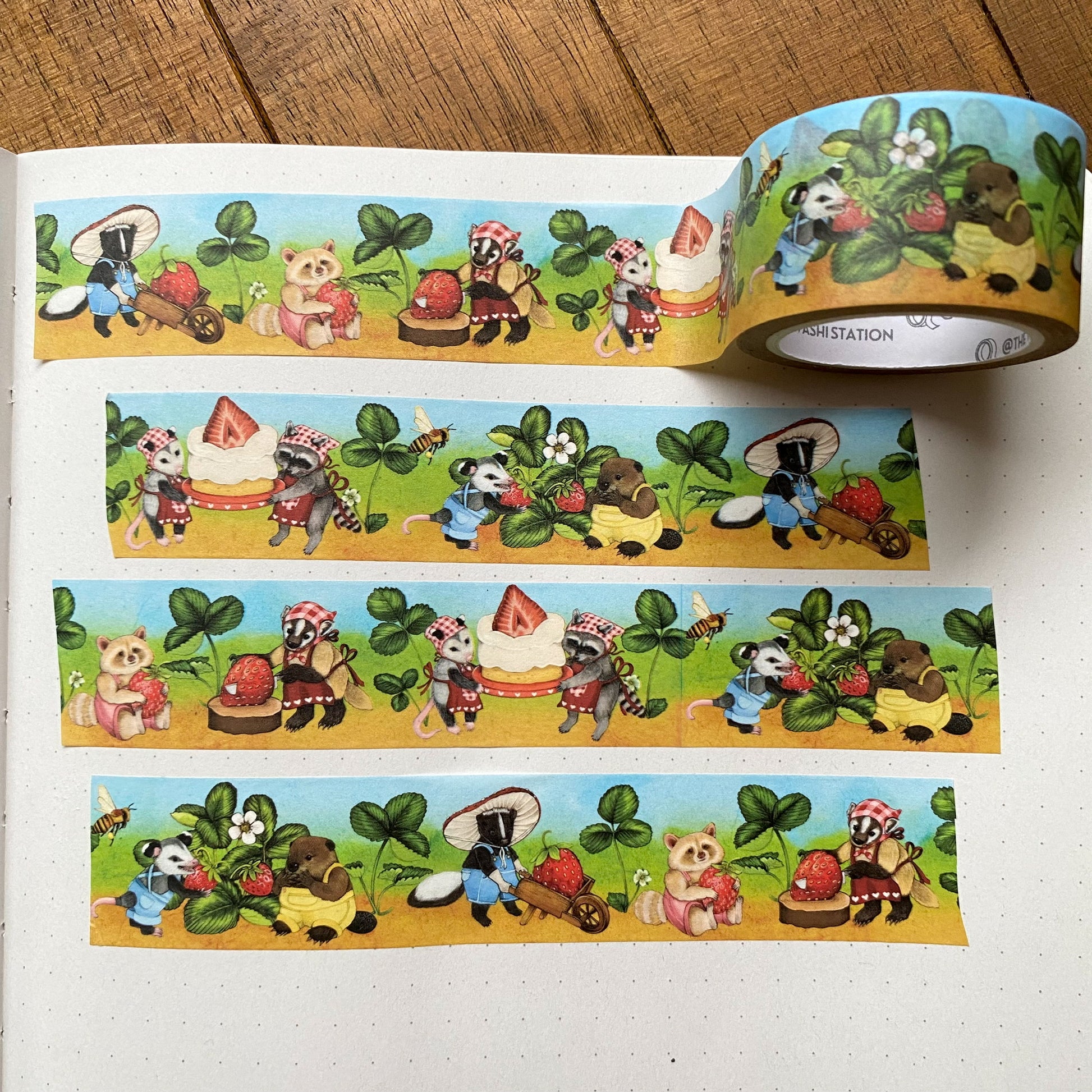 Washi tape of a farm scene with an opossum picking strawberries that a skunk takes in a wheelbarrow to a badger, who cuts the strawberry. Then there are a raccoon and opossum holding a cake. The washi tape measures 25mm and is swatched on a notebook.