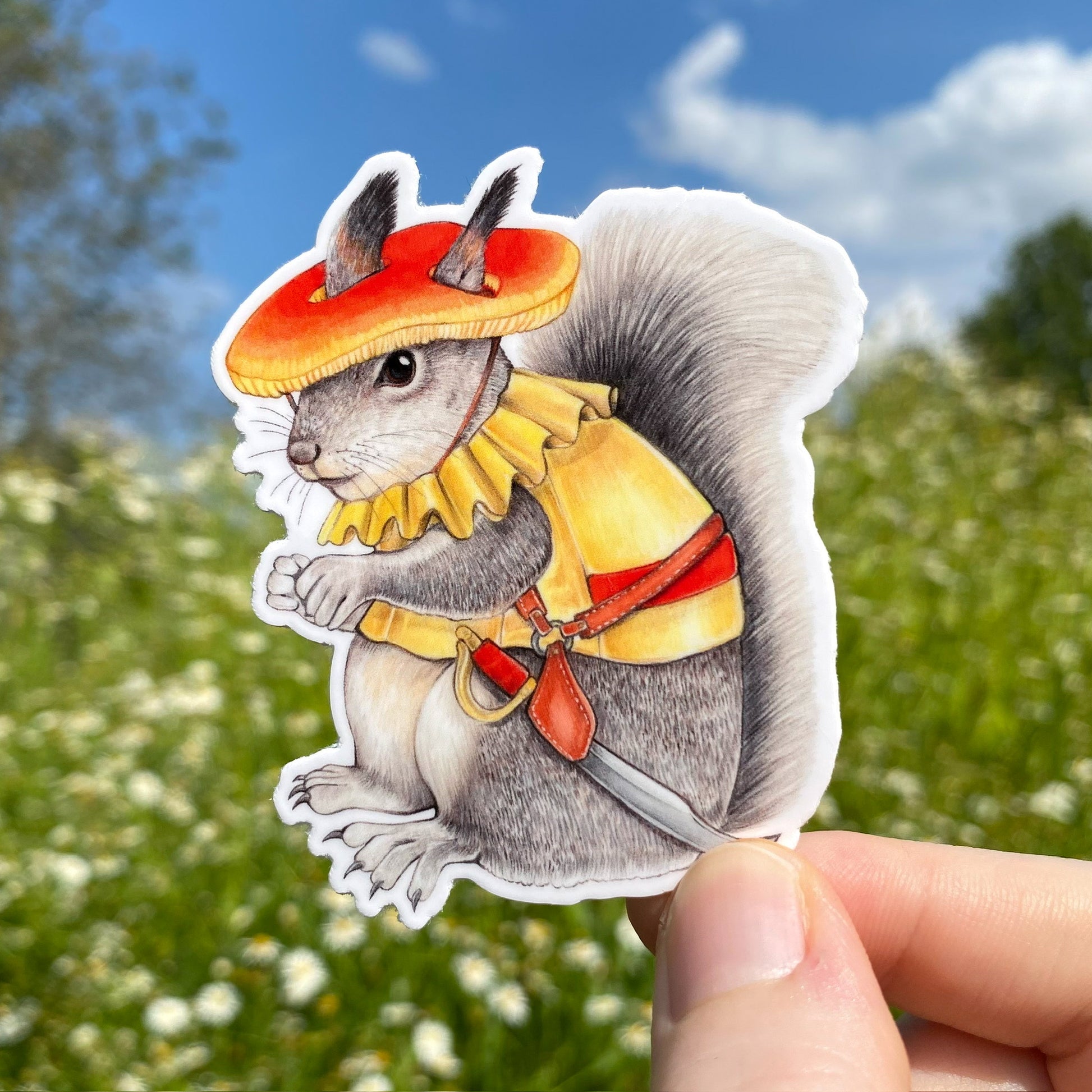 A hand holding a weatherproof vinyl sticker of an Abert's squirrel with a red amanita hat and yellow vest. He has a sword holstered to his hip.