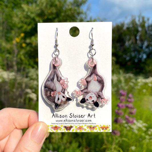 A hand holding a pair of acrylic earrings of two baby opossums hanging upside down.