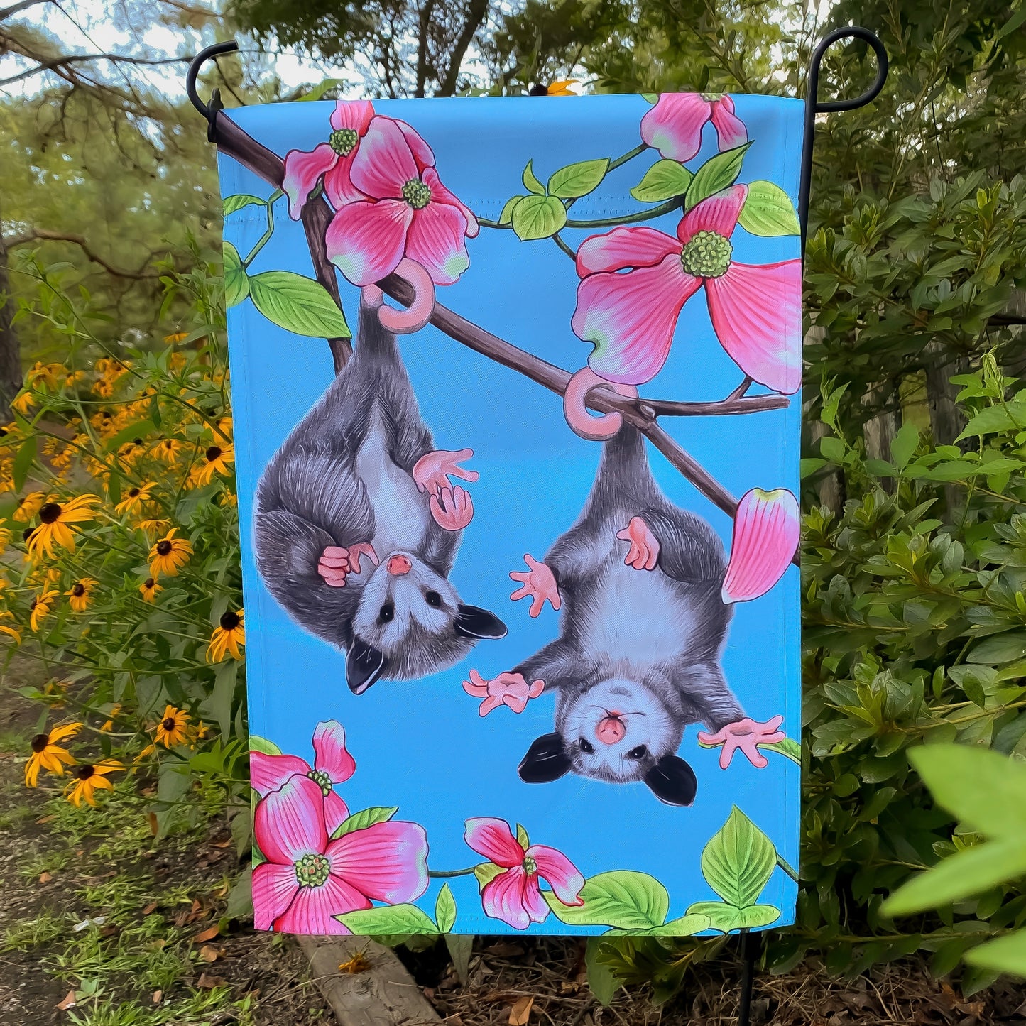A garden flag of two baby opossums hanging from a pink flowering dogwood tree.