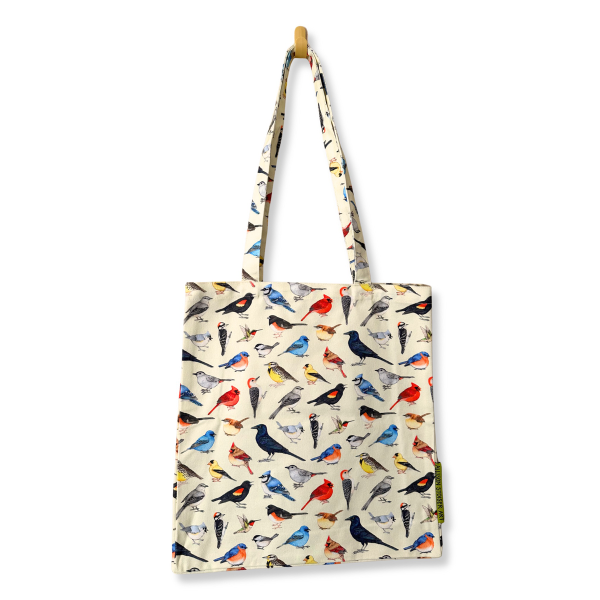 A canvas tote bag with a backyard bird pattern on a cream background, hanging from a wooden peg on a white wall.