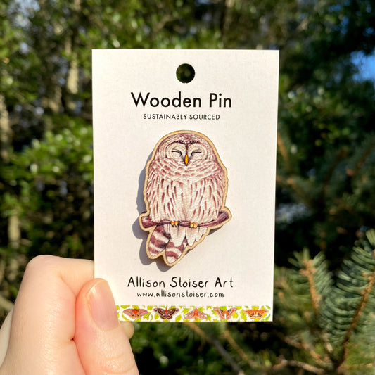 A hand holding a wooden pin with an illustration of a barred owl.