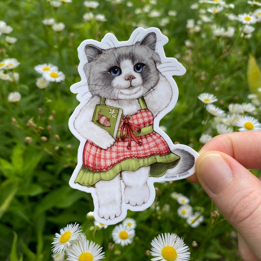 Sticker of a cat in a dress holding a book held in front of greenery with flowers