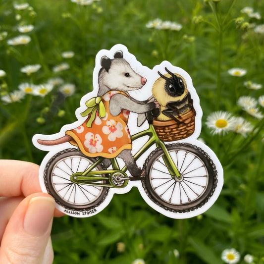 Sticker of an opossum in a dress riding a bike with a bee in a basket against a natural background.