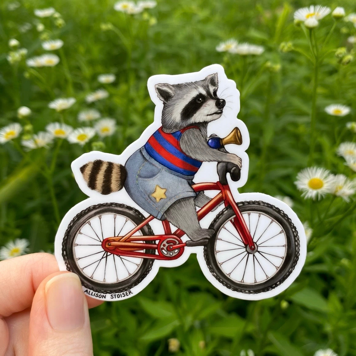 Sticker of a raccoon on a bicycle held in front of a green field with flowers