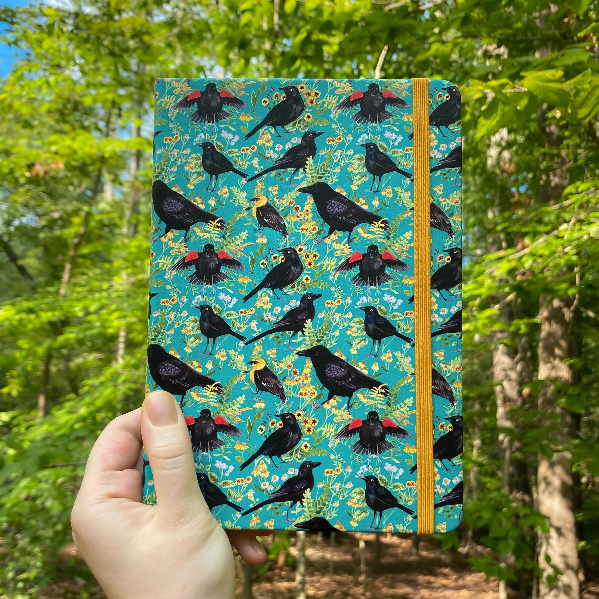 Notebook with black bird pattern held in front of a forest background