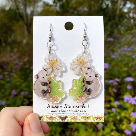 Opossum and bloodroot earrings on a card held by a hand with a natural background