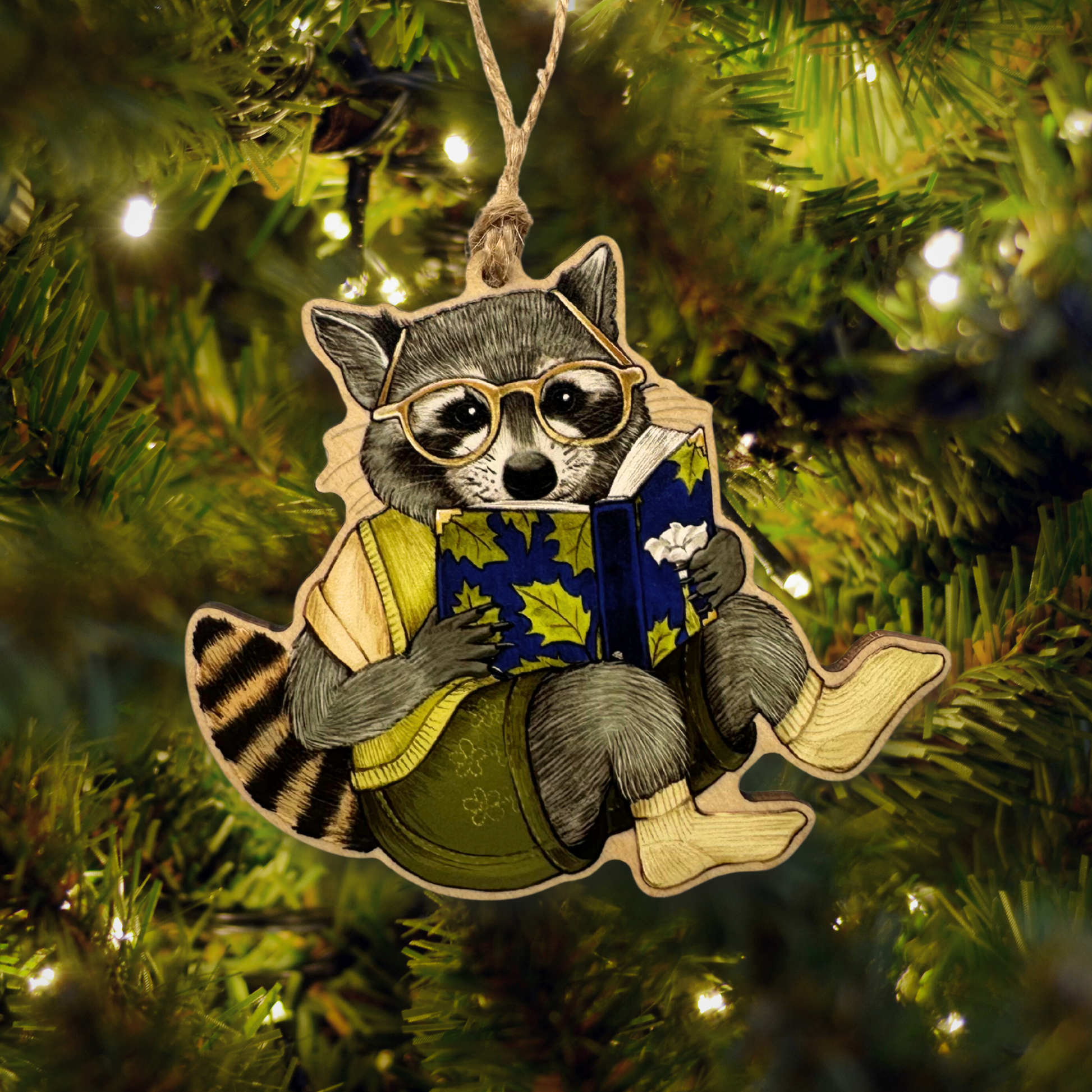 Decorative ornament of a raccoon wearing glasses and reading a book, hanging on a Christmas tree.