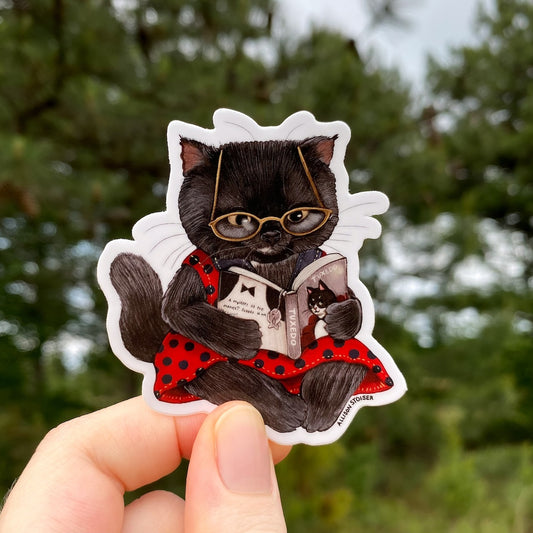 Sticker of a black cat wearing glasses and a red dress holding a book, held in front of a blurred green background.