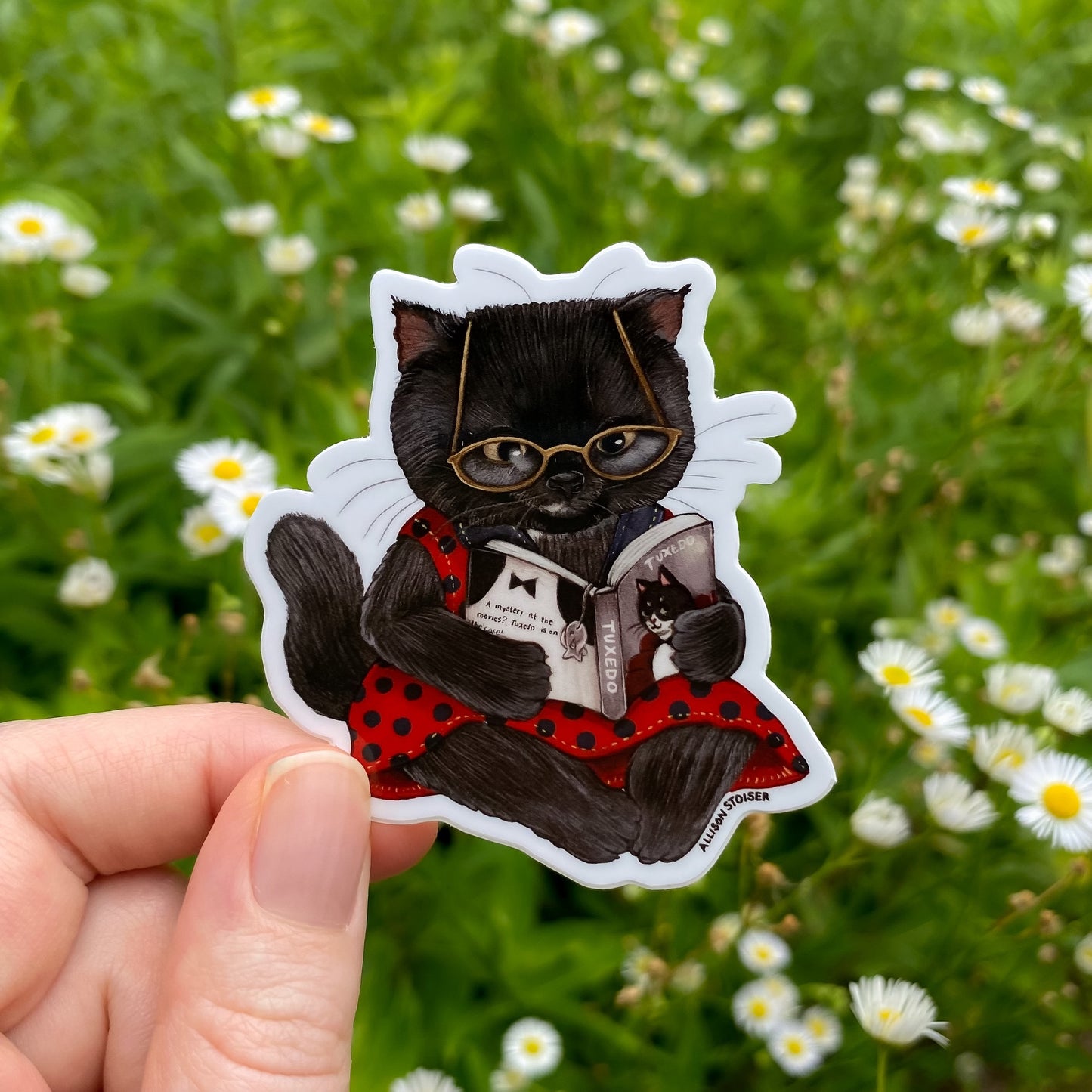 Sticker of a black cat wearing glasses and a red outfit held in front of a green field with flowers.