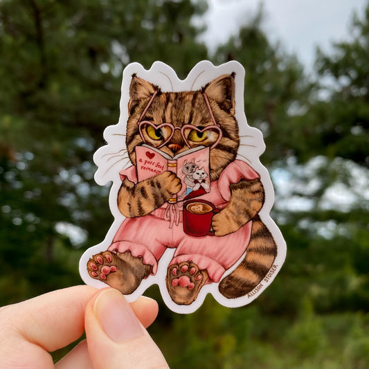 Sticker of a brown tabby cat reading a book with heart-shaped glasses held by a hand against a blurred green background.