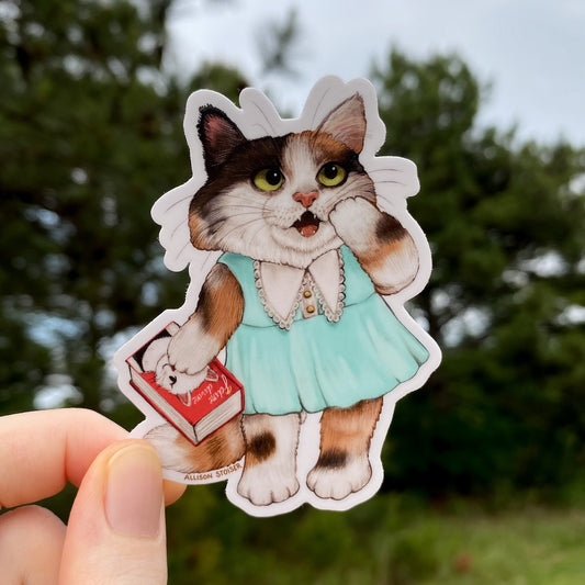 Sticker of a calico cat in a blue dress held by a hand with a blurred natural background