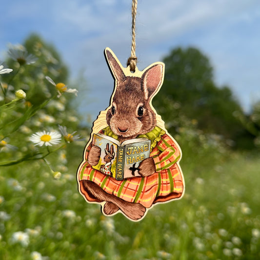 A hanging wooden ornament of a rabbit in a plaid dress reading a book, photographed outdoors against a blurred meadow and blue sky background.