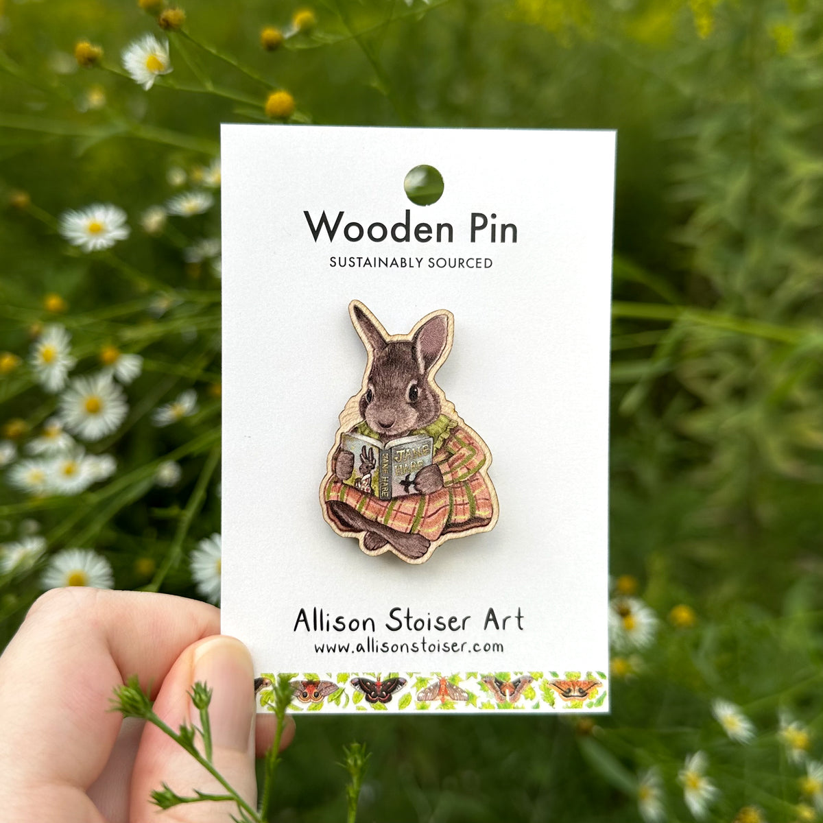 A wooden pin featuring a rabbit reading a book, mounted on a white display card held by a hand against a blurred background of green foliage and asters.