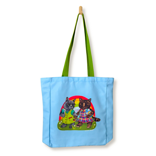 A light blue canvas tote bag with bright green shoulder straps, featuring a print of two cats in dresses on the front, hanging from a wooden peg against a white background.