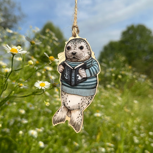 A hanging ornament of a seal wearing a striped sweater and holding a book, photographed outdoors against a blurred field of white flowers and green grass under a blue sky.