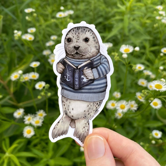 Sticker of a seal wearing a sweater holding a book, held in front of a natural background with flowers.
