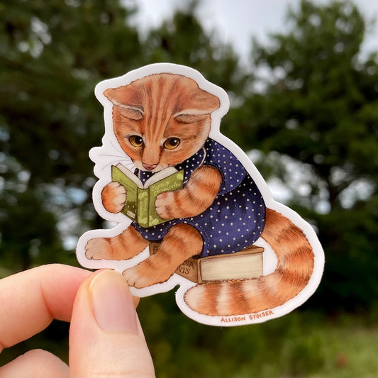 Sticker of an orange tabby cat reading a book held by a hand with a blurred green background