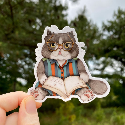 Sticker of a cat wearing glasses and reading a book, held against a blurred green outdoor background.