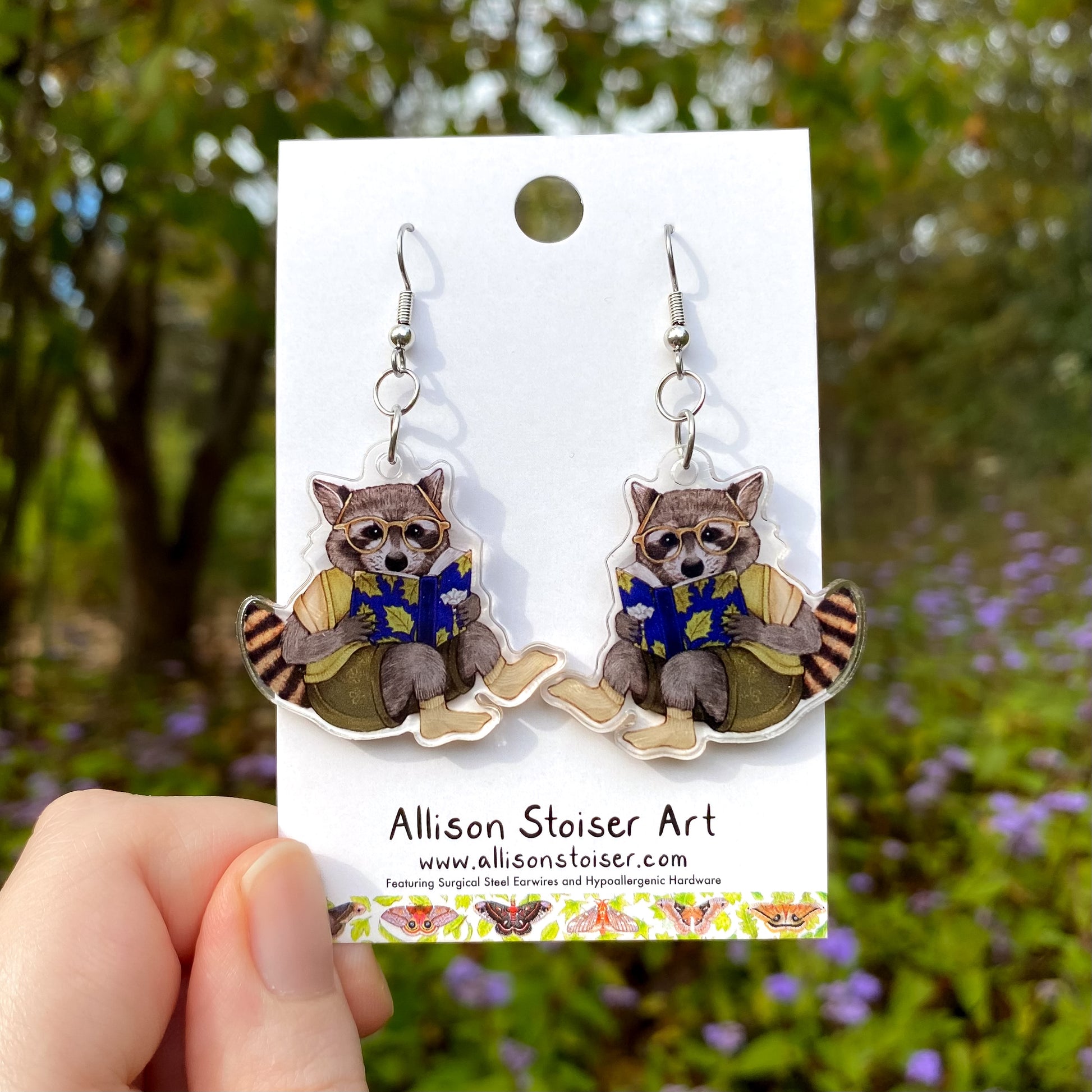 Raccoon earrings on a card held in front of a natural background