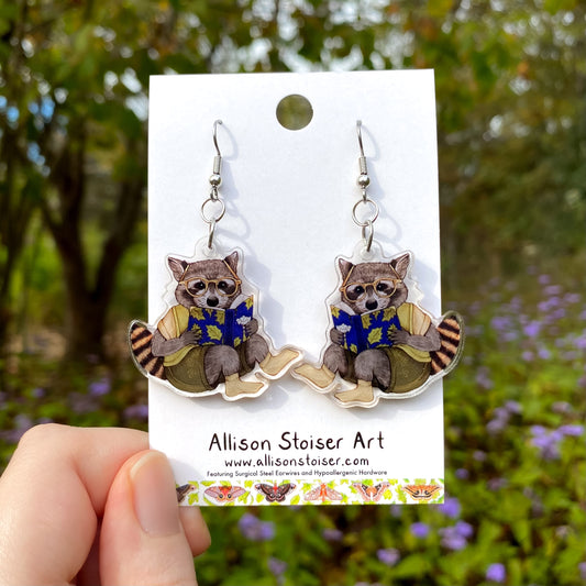 Raccoon earrings on a card held in front of a natural background