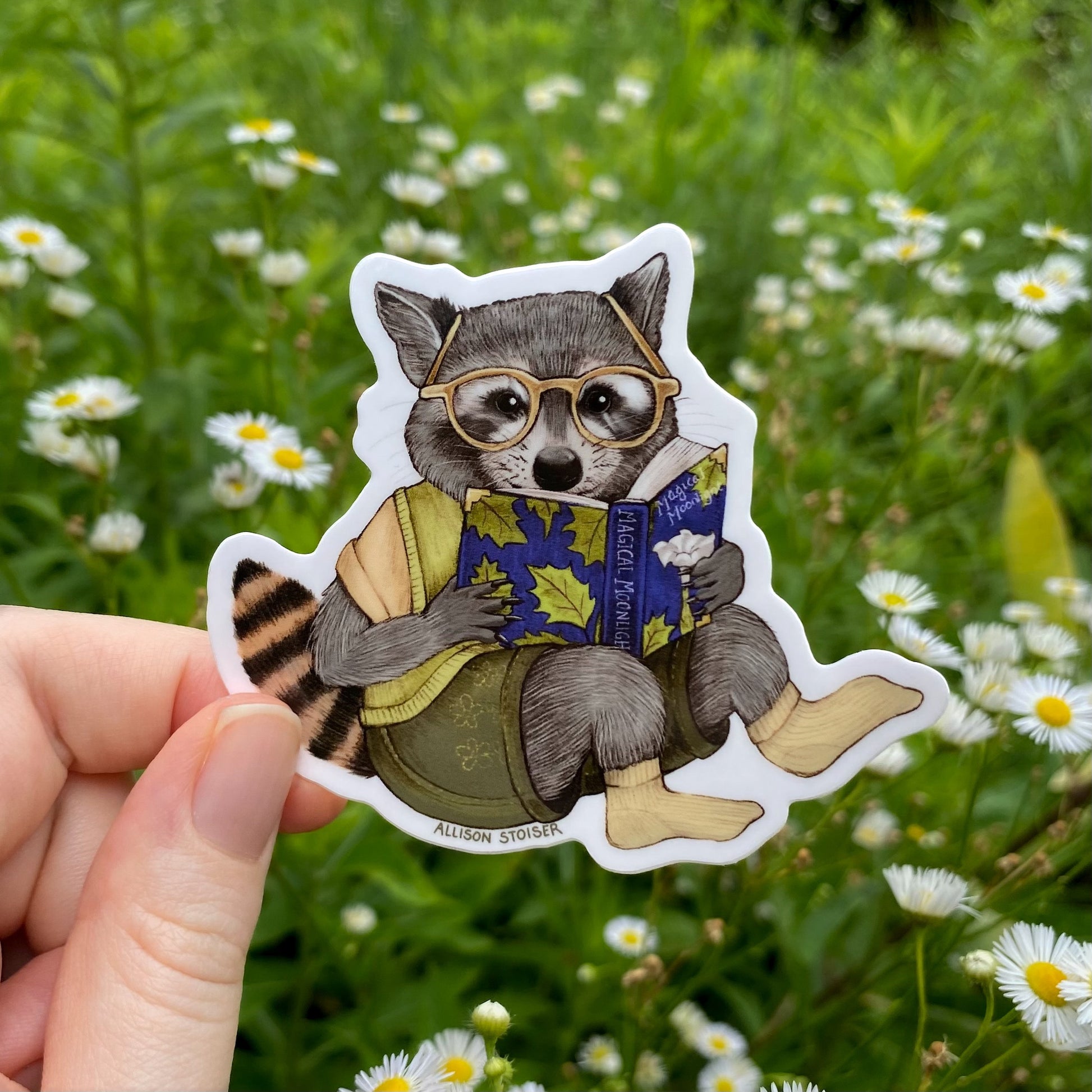 Sticker of a raccoon wearing glasses and reading a book held in front of a natural background with flowers.