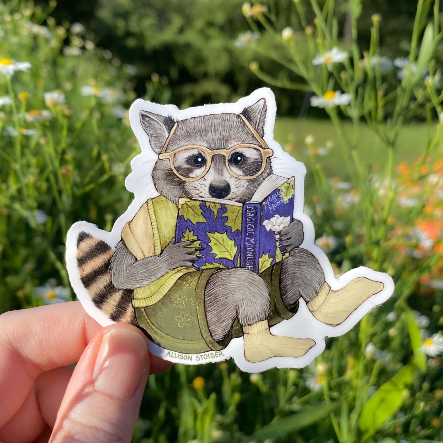 Sticker of a raccoon wearing glasses and reading a book held in front of a natural background with flowers and grass.